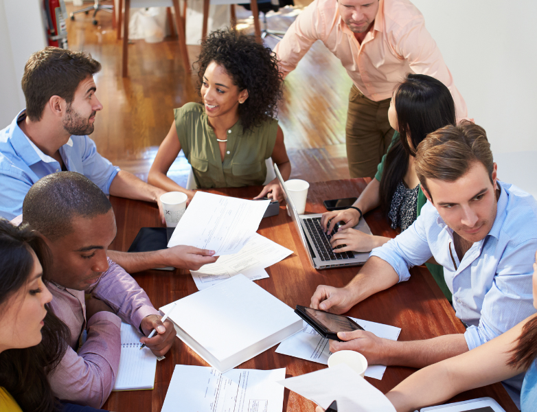 A diverse group of eight people sit around a wooden table in a modern office, collaborating on documents and laptops, while engaged in discussion. Papers, notebooks, coffee cups, and digital devices are scattered on the table.