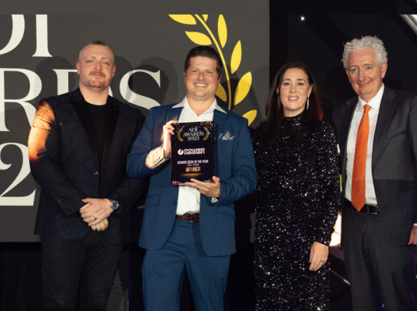 Four people in formal attire stand together at an awards event. The person second from the left is holding a plaque that reads SQI Awards 2023 WINNER. A decorative backdrop and lighting are visible behind them.
