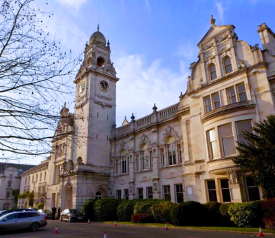 A historic stone building with ornate architecture, arched windows, and a clock tower stands under a blue sky, bordered by trees, bushes, and parked cars. This is Kingston County Hall in Surrey.