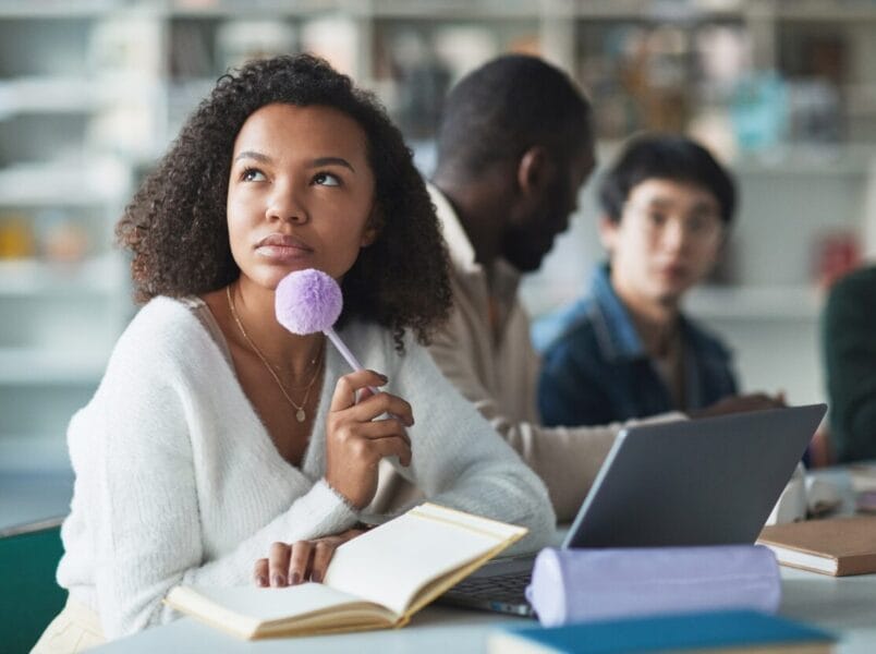 A young woman sits at a table with a notebook and laptop, holding a fluffy pen and looking thoughtfully upward—perhaps considering selecting an ITSM tool. Two others are blurred in the background, suggesting a group study setting.
