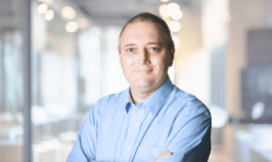 A smiling John Noctor, SDI's Chief Customer Success Officer, dressed in a professional suit and tie.