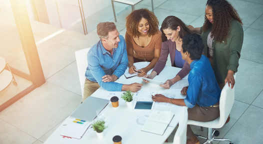 A diverse group of five people smiling and collaborating around a table with documents, coffee cups, and digital devices in a bright, modern office setting.