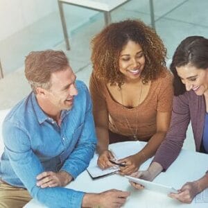 Five people sit around a table, smiling and collaborating on a project with papers, a tablet, and a notebook in a bright, modern office space. Sunlight streams in from large windows.