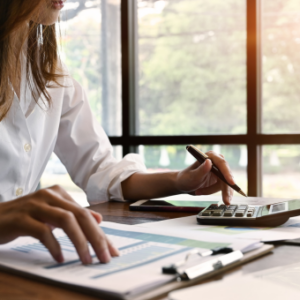 A person in a white shirt working at a desk, using a calculator and reviewing documents with charts and graphs, proving ROI in ITSM as productivity is transformed, with large windows and greenery visible in the background.
