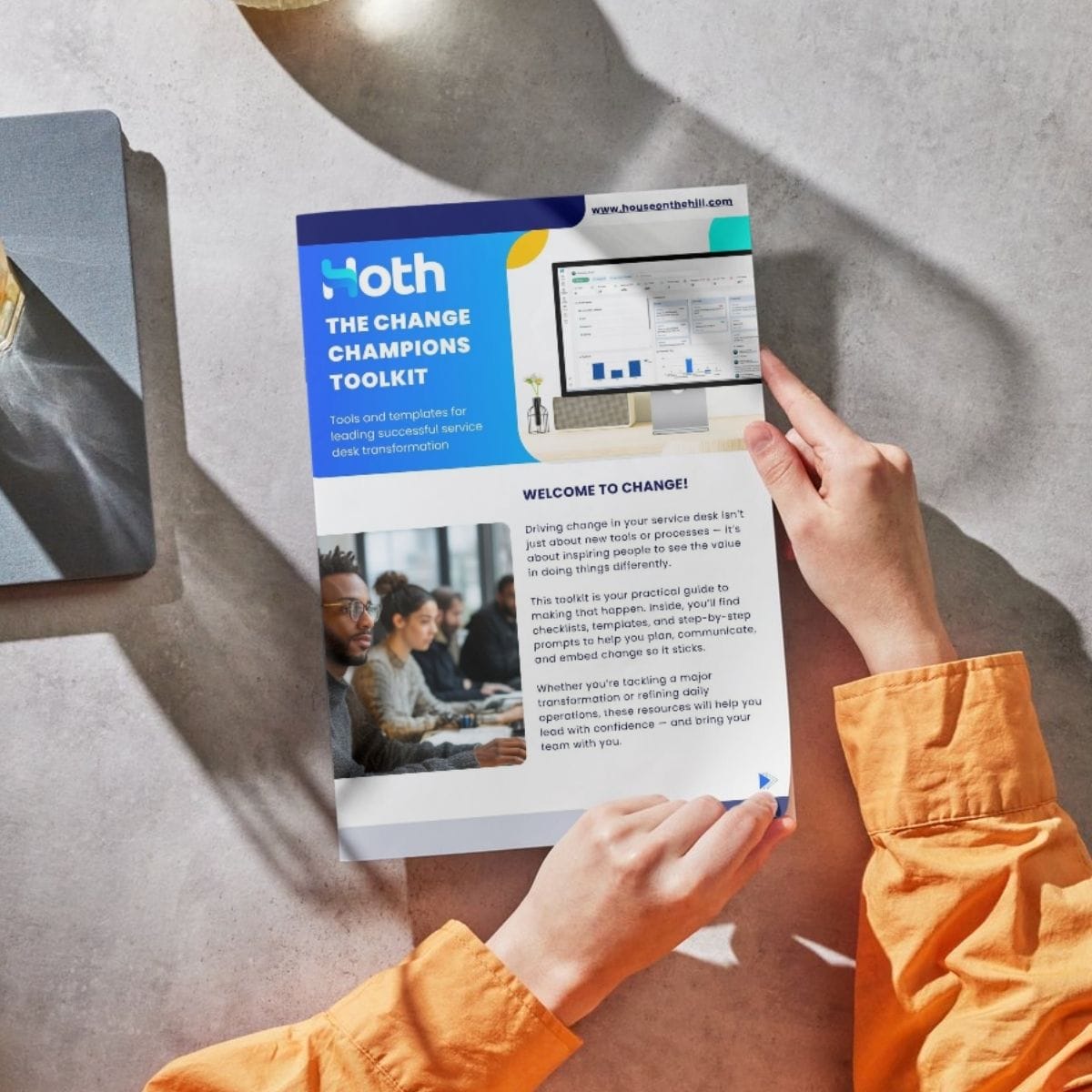 A person in an orange shirt holds a printed guide titled The Change Champions Toolkit at a table; a laptop and drink sit nearby. The flyer highlights Service Desk Change Management tips, images, and a website link for leading successful transformation.