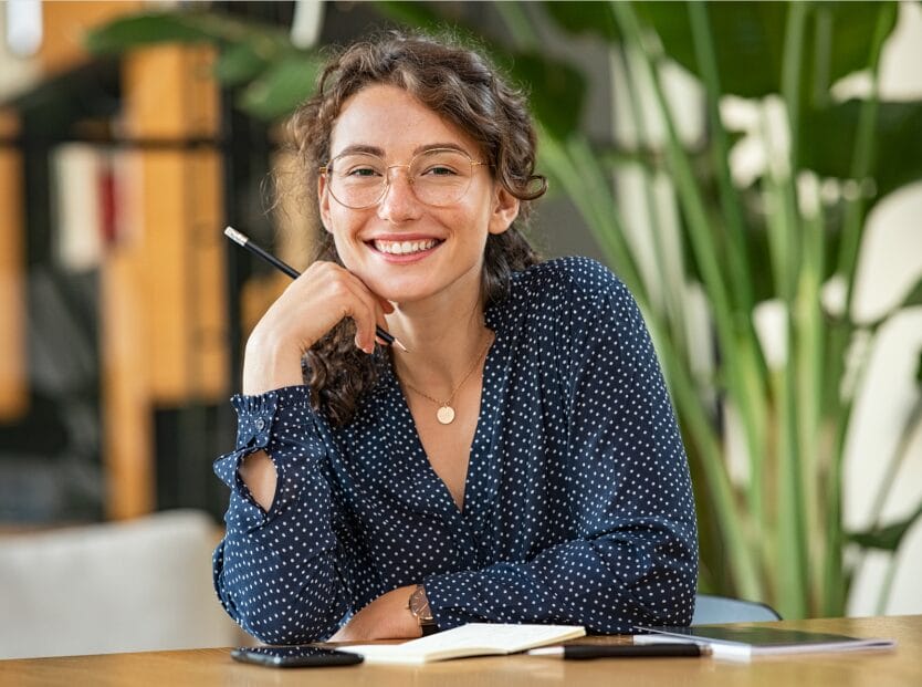 A thoughtfully posed individual rests their chin on their hand, seated at a desk with a notepad and a phone, surrounded by greenery.