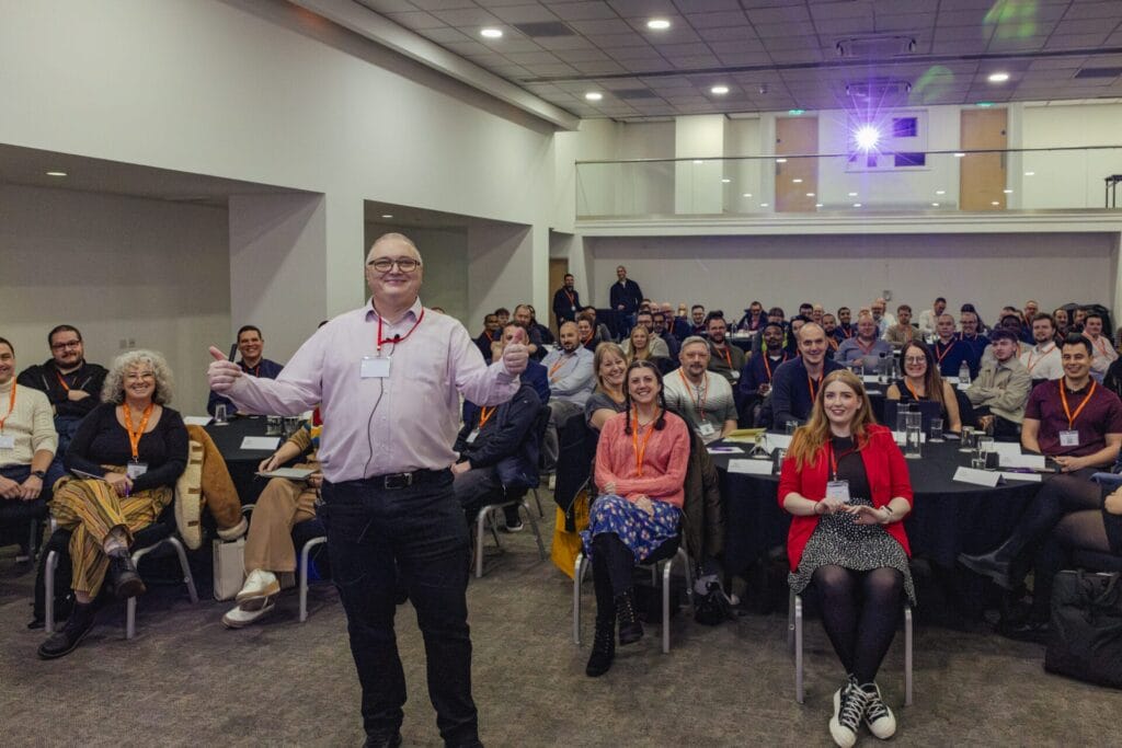 A man in a light pink shirt stands smiling with both thumbs up in front of a large seated audience at a Service Desk Continual Improvement conference. The audience members are smiling and looking towards the camera.