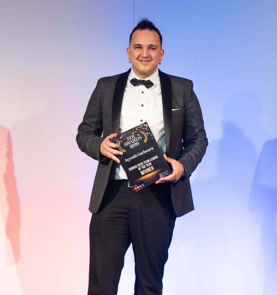 A man in a tuxedo stands on stage, smiling and holding an award plaque. The background features a gradient of white to blue, and stage lighting is visible in the foreground.