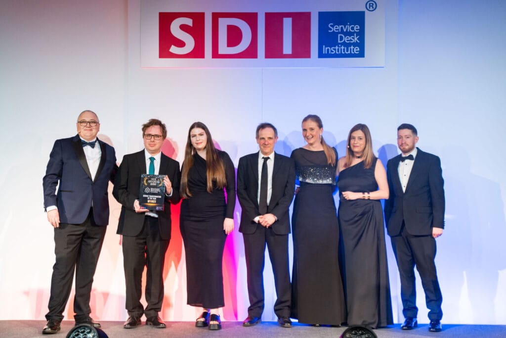 Seven people in formal attire stand on stage under a Service Desk Institute sign. One person holds an award plaque, and the group is smiling, posing for a photo at an event.