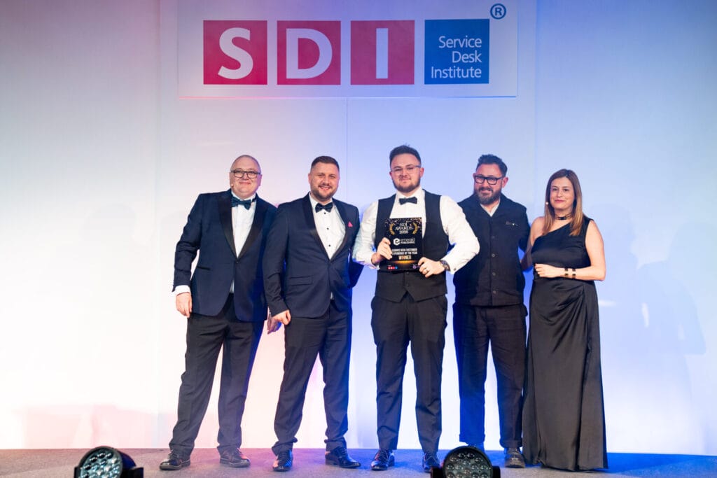 Five people in formal attire stand on stage under a Service Desk Institute (SDI) sign. One person in the centre holds an award, and they are all smiling for the photo.