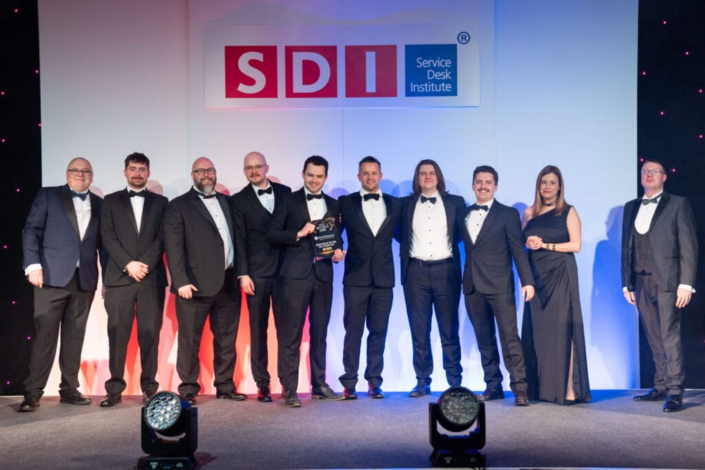 A group of ten people in formal attire stand on stage, smiling, with one person holding an award. Behind them is a large SDI (Service Desk Institute) sign on a white and blue-lit backdrop.