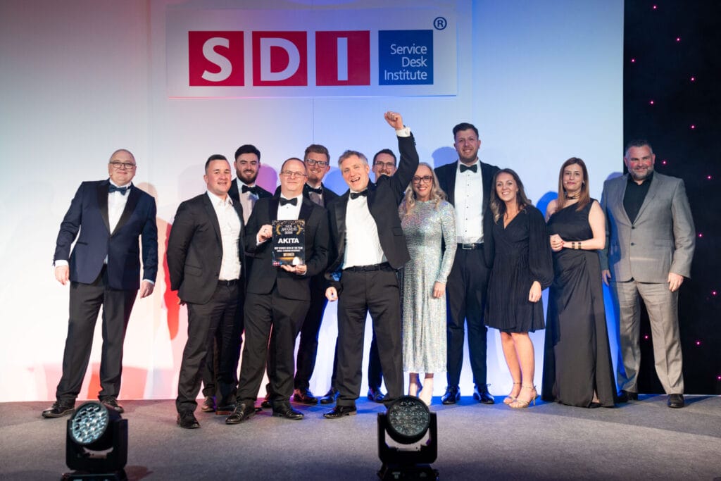 A group of people in formal attire stand on stage under an SDI (Service Desk Institute) sign, smiling and celebrating as one man holds up a trophy, with others applauding and posing for the photo.