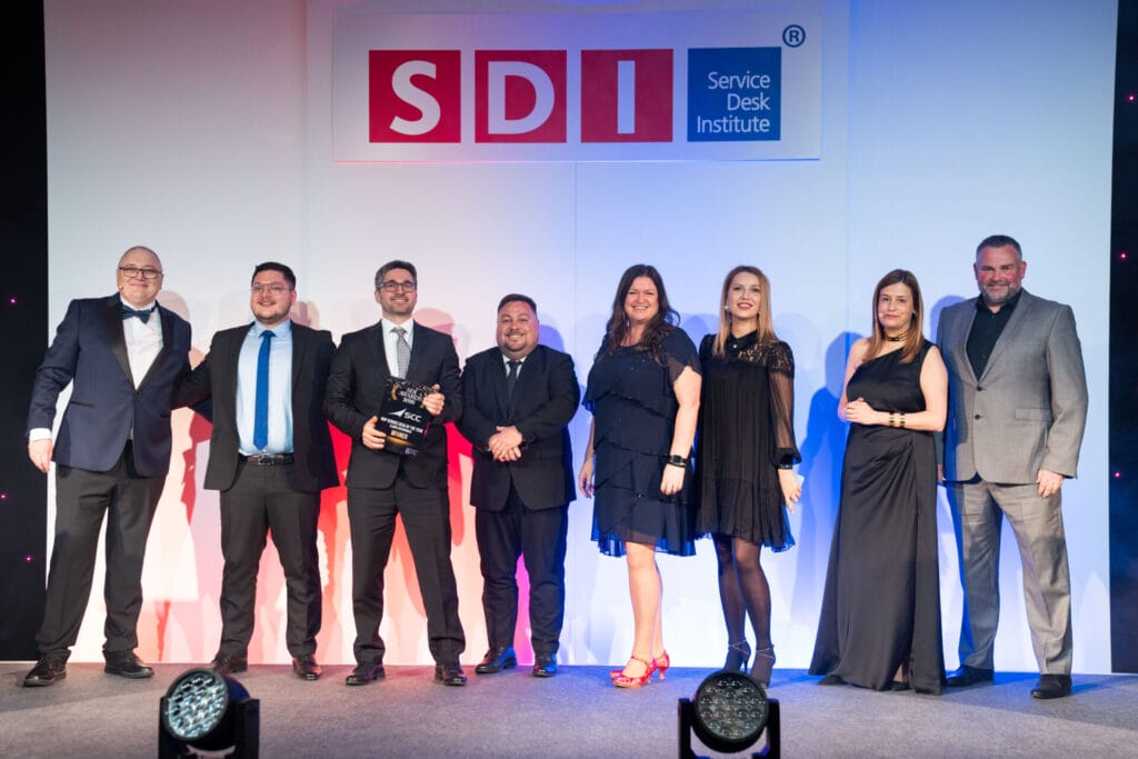 Eight people dressed in formal attire stand on stage under a Service Desk Institute (SDI) sign, smiling and posing together at an awards event. One person holds an award.