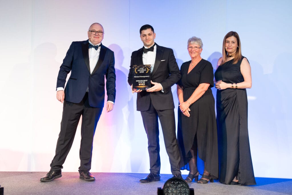 Four people stand on a stage in formal attire, smiling. The man second from the left holds a glass award. The group appears to be at an awards ceremony with a plain white and blue-lit backdrop.