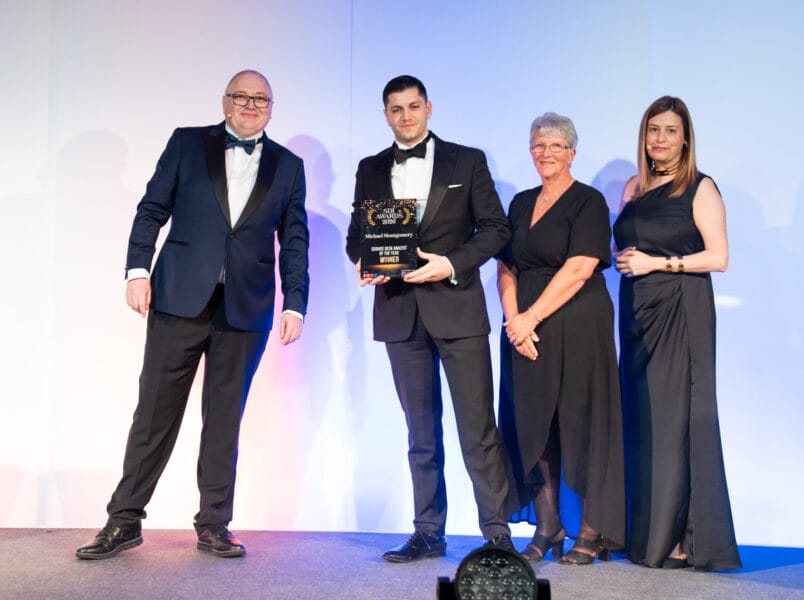 Four people stand on a stage in formal attire, smiling. The man second from the left holds a glass award. The group appears to be at an awards ceremony with a plain white and blue-lit backdrop.