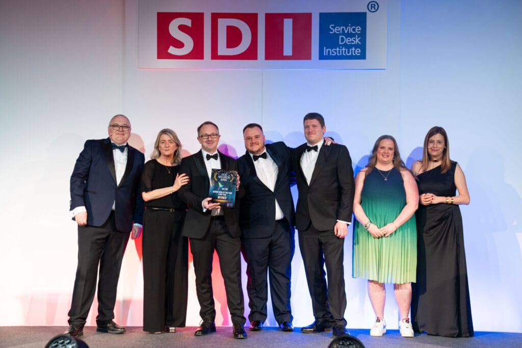 Seven people in formal attire stand onstage under an SDI (Service Desk Institute) sign. The group smiles as one person holds an award plaque, indicating an achievement or recognition event.