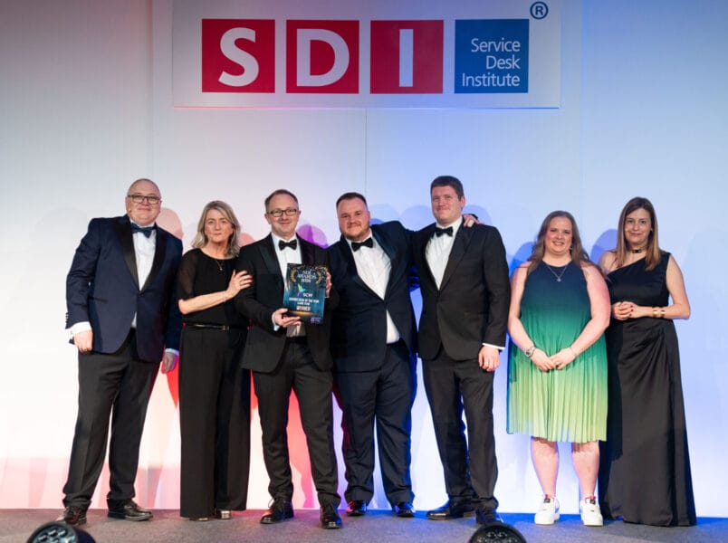Seven people in formal attire stand onstage under an SDI (Service Desk Institute) sign. The group smiles as one person holds an award plaque, indicating an achievement or recognition event.