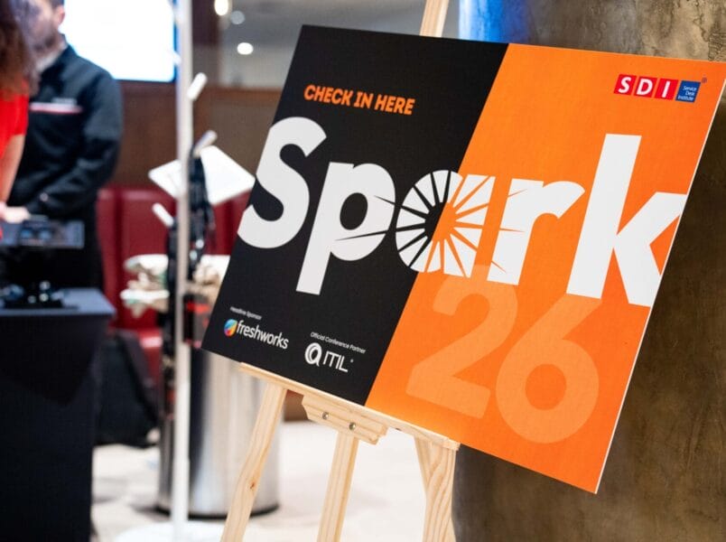An event sign on an easel reads Check in here SDI Conference: Spark 27 with logos for SDI, Freshworks, and ATL. People stand in the background, slightly out of focus.