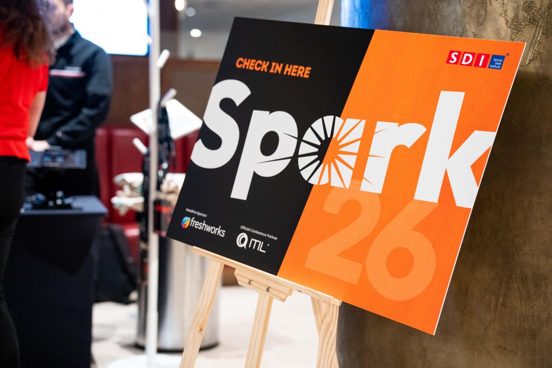 An event sign on an easel reads Check in here SDI Conference: Spark 27 with logos for SDI, Freshworks, and ATL. People stand in the background, slightly out of focus.