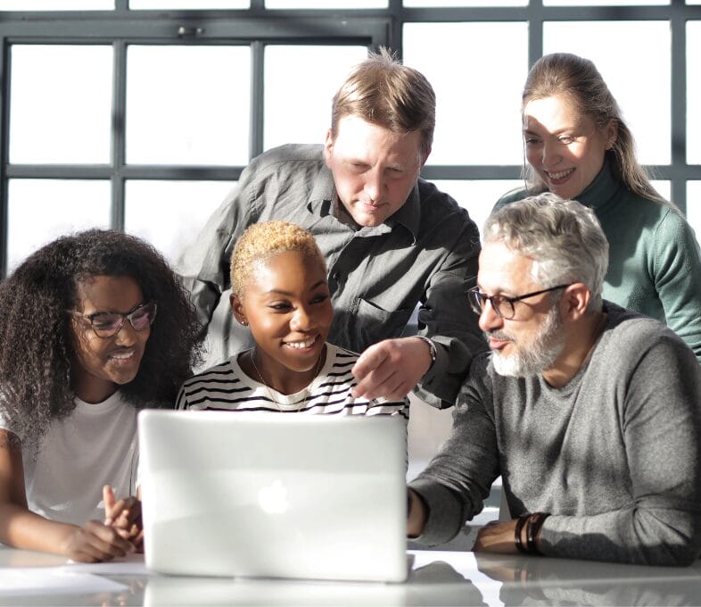 Five people sit and stand around a table, smiling and looking at a laptop screen together in a bright room with large windows, reflecting a team focused on building IT resilience that compounds.