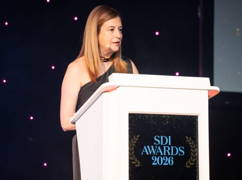 ITIL presenter, Chaido, hosting the awards ceremony in a black dress stands at a white lectern labelled SDI Awards 2026, addressing the audience from the stage as purple lights shine in the background.