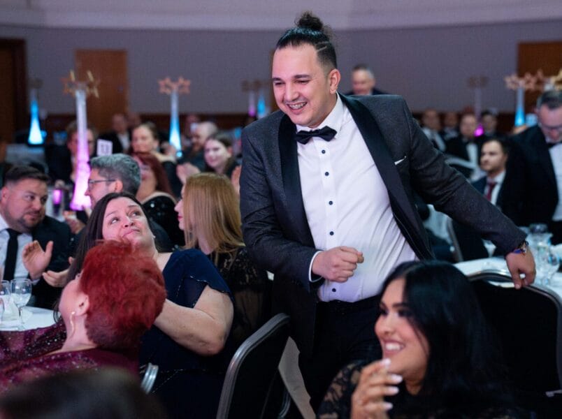 A man in a dinner jacket smiles and stands up between tables at the SDI Awards 2026, while people around him applaud and look at him with happy expressions. The room is filled with guests seated at decorated tables.
