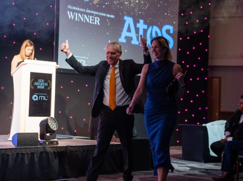 A man and woman walk excitedly to the stage at an awards ceremony, with the man giving two thumbs up. Behind them, a woman stands at a lectern and a screen displays WINNER and the Atos logo.