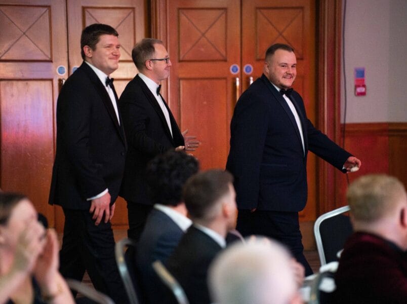 Three men in black dinner jackets walk side by side through a room full of seated people, heading towards the front. Wooden doors are visible in the background. The setting appears to be a formal event or ceremony.