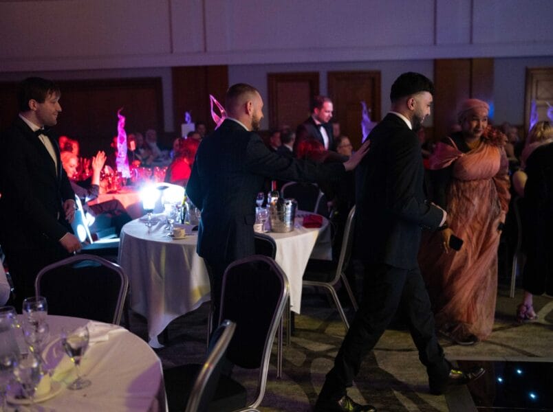 A group of people in formal attire walk past round tables at a dimly lit event or banquet, with guests seated and colourful lights illuminating the background.