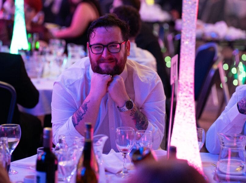 A man with glasses and tattoos on his arms sits at a formal event table, resting his chin on his hands and smiling. The room is decorated with colourful lights and there are glasses and bottles on the table.