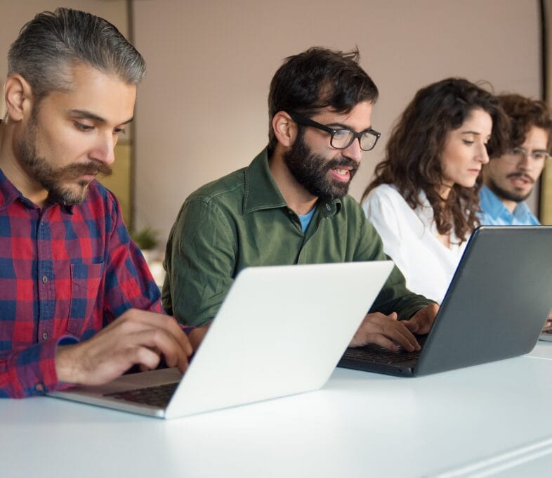 Four people sit in a row at a white table, focused on their laptops as they collaborate on an AI-ready ServiceOps roadmap. Two men in front wear checked red and green shirts; two others with curly hair work in the background.