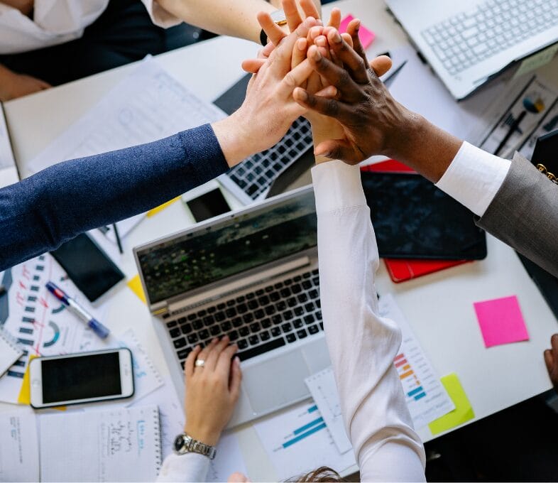 A group of people in business attire gather around a desk with laptops, documents, and charts, raising their hands together in a high-five gesture, symbolising teamwork and collaboration.