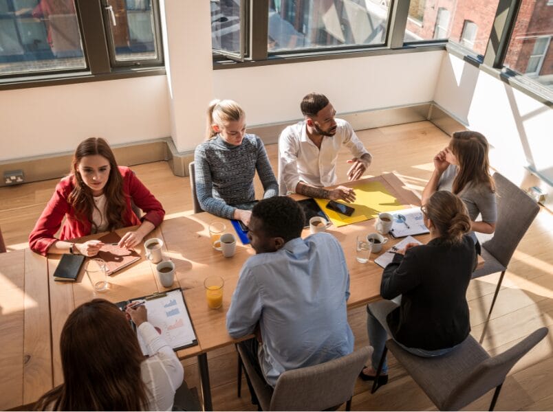 A group of seven people sit around a table in a bright office, engaged in discussion during a high performing teams workshop. Papers, notebooks, coffee cups, and a tablet are on the table as sunlight streams through large windows.