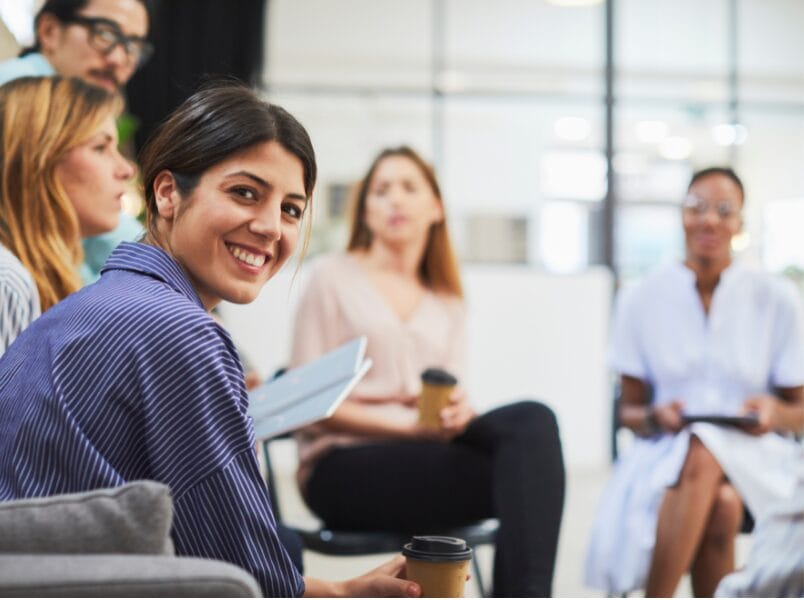 A woman in a striped shirt smiles at the camera whilst sitting with a group during a high-performing teams workshop in a bright, modern office. Others hold coffee cups and chat in the background.
