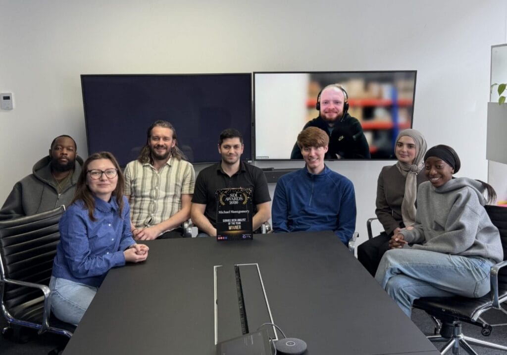 The Wanstor's Leonard Cheshire team - Eight people pose in a meeting room, seven seated around a table and one appearing on a large screen via video call. The group smiles, and th SDI Service Desk Analyst of the Year 2026 Award trophy is displayed on the table.