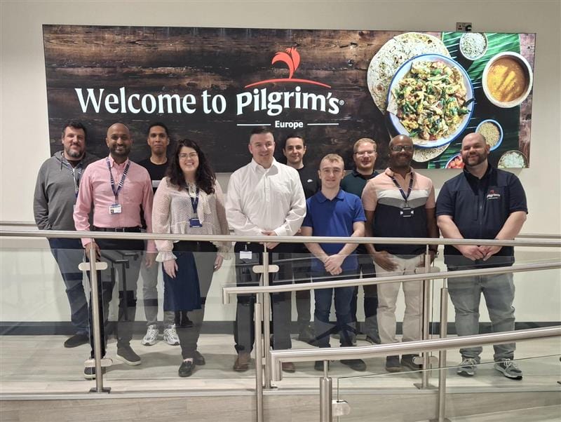 Ten people stand smiling in front of a Welcome to Pilgrims Europe sign adorned with food images, proudly posing near a railing in a brightly lit indoor setting, celebrating their recent Service Desk Certification achievement.