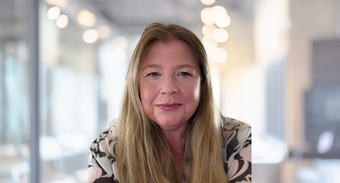 A woman with long blonde hair and a patterned blouse smiles softly at the camera in a bright, blurred office setting.