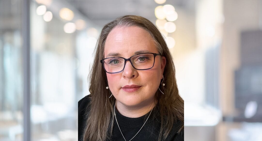 Sophie Hussey, a person with long brown hair, glasses, and earrings, wearing a black top, poses indoors with a blurred office background and soft lighting.