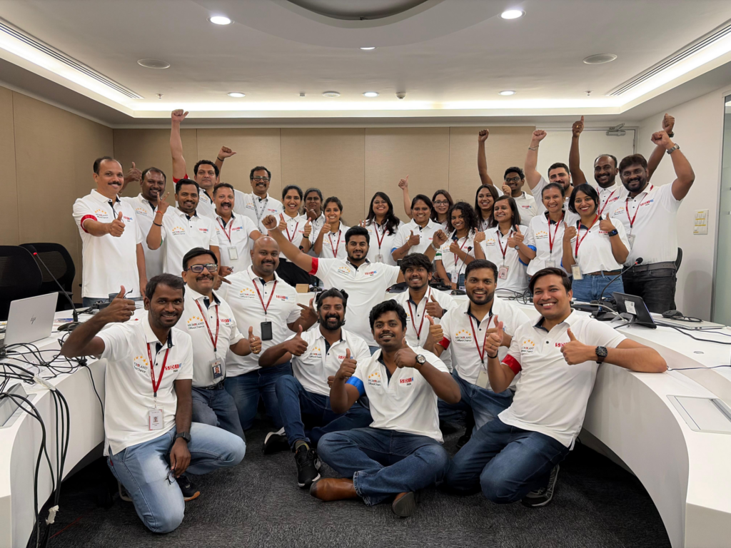 A group of people in white shirts and jeans pose together in an office, smiling and giving thumbs up. They appear happy and excited, standing and sitting around conference tables with laptops.