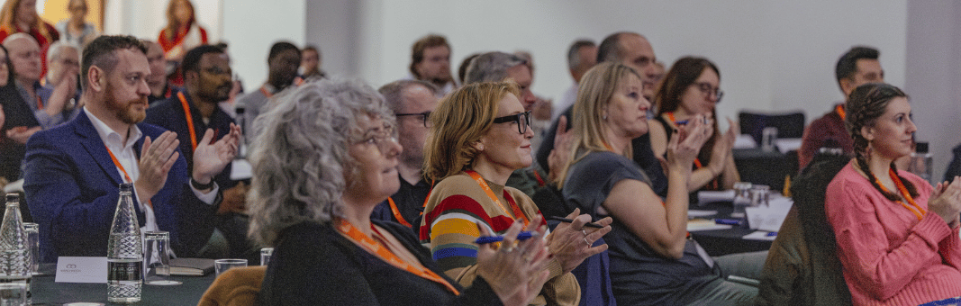 A diverse group of attendees sitting at tables in a conference room, applauding while looking towards a presentation.