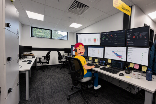 An illustration of a cartoon person wearing a headset sits at a desk in an IT office, facing a wall of monitors displaying system health and security dashboards.